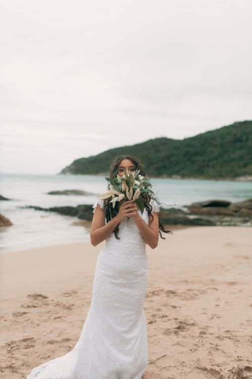 Free A bride in a white dress stands on a beach holding a bouquet, with the ocean and hills in the background. Stock Photo