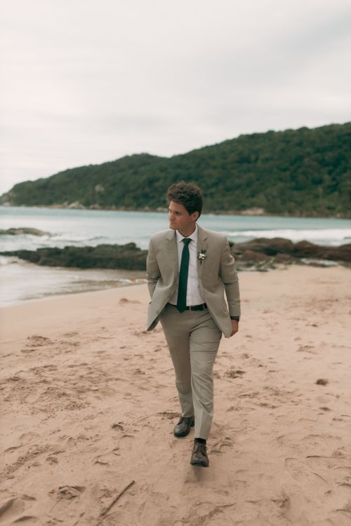 Free Man in a beige suit strolling along a sandy beach with lush hills in the background. Stock Photo