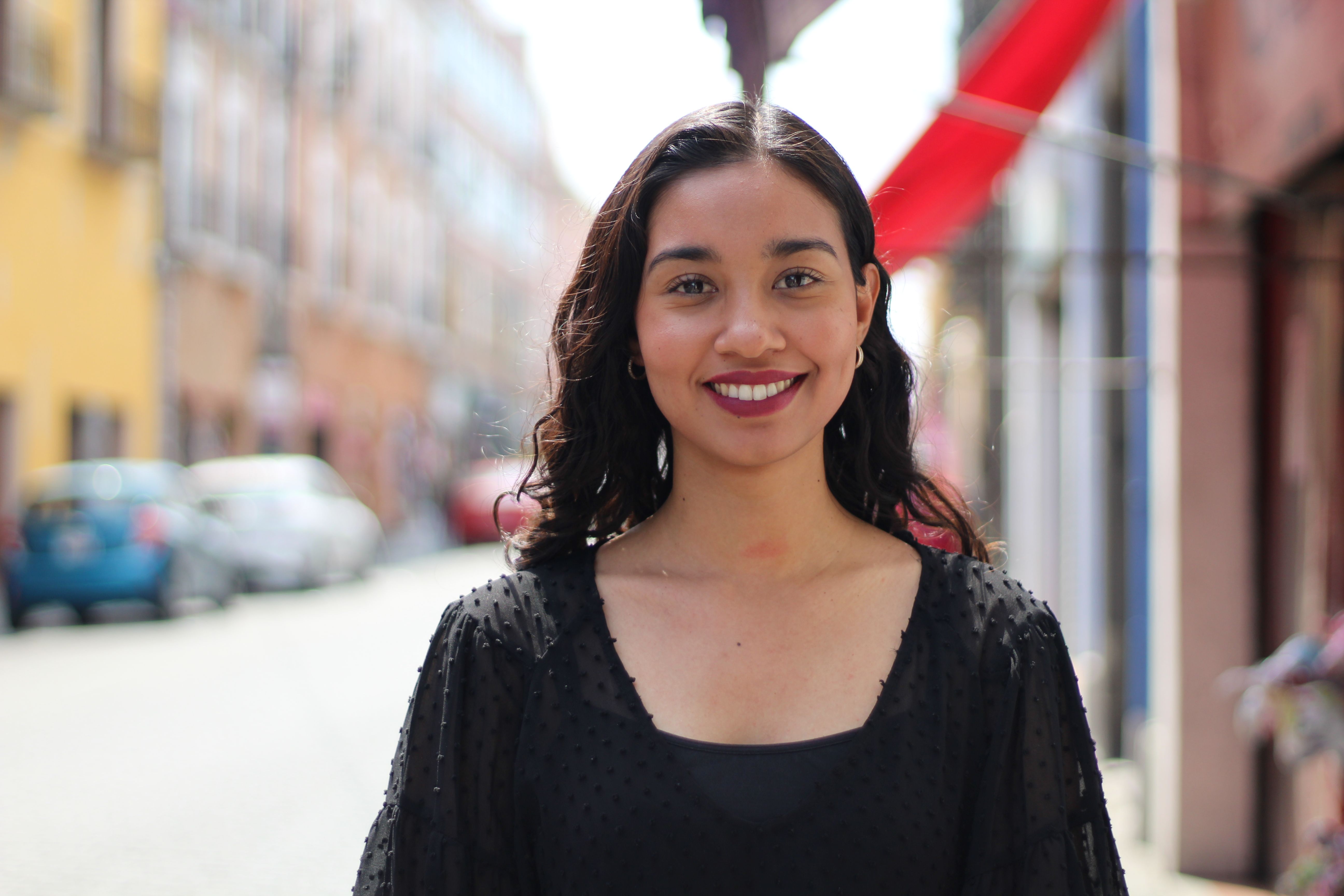 A woman in a black top smiles warmly while standing on a vibrant city street.