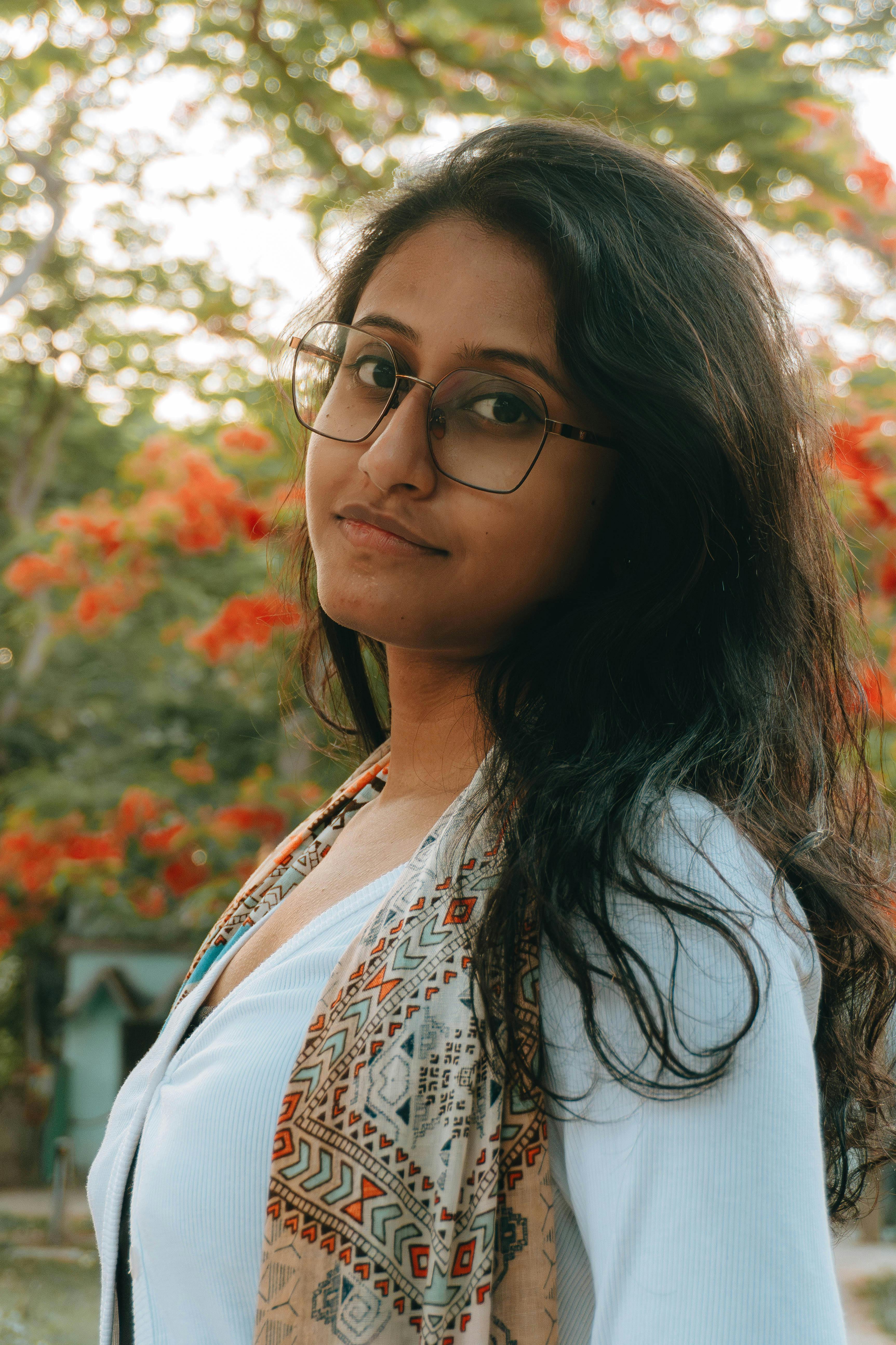 A woman with glasses poses outdoors against a blooming backdrop, capturing a warm autumn vibe.