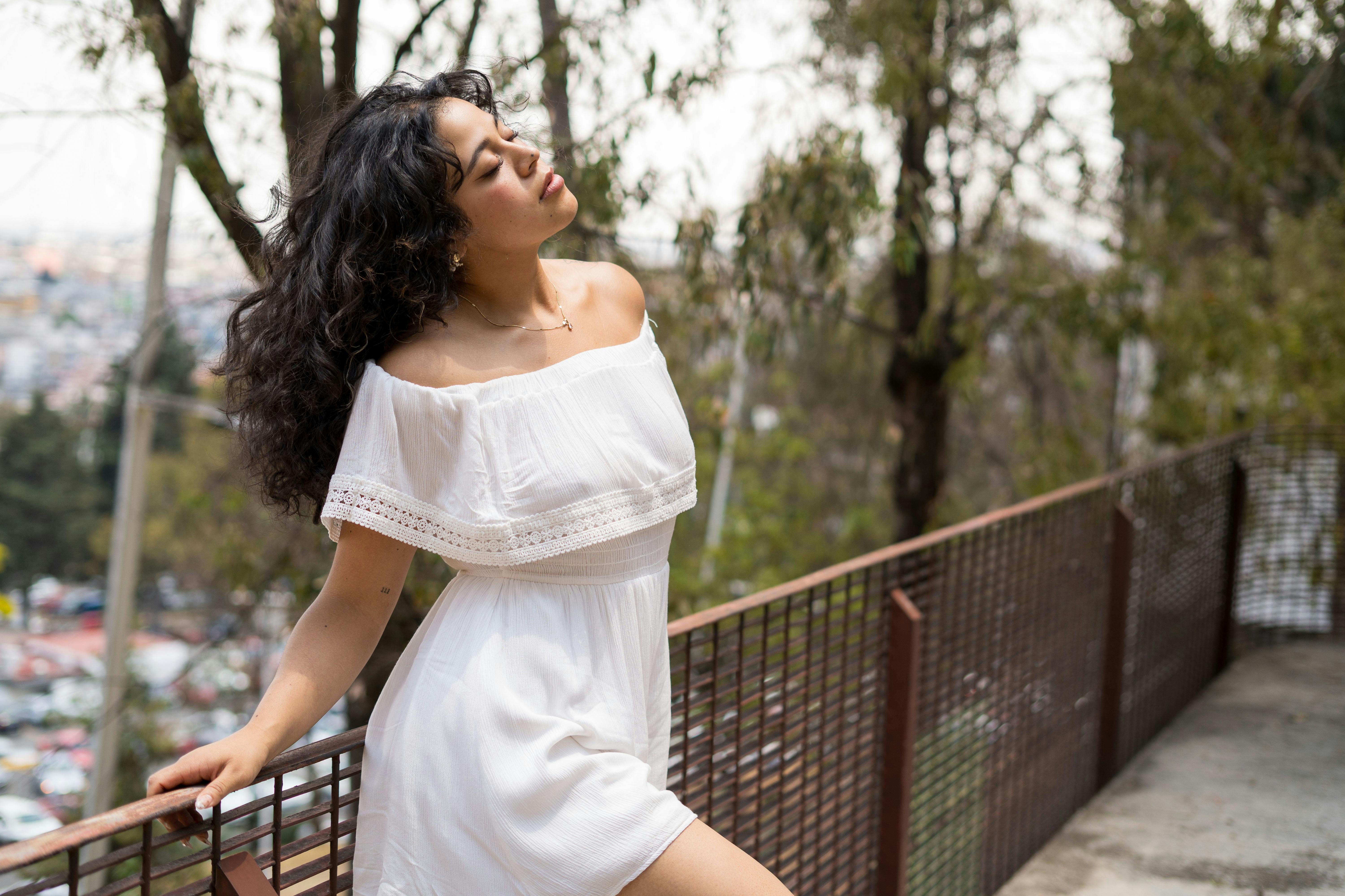 A young woman in a white dress leans on a railing in an outdoor setting, enjoying nature.