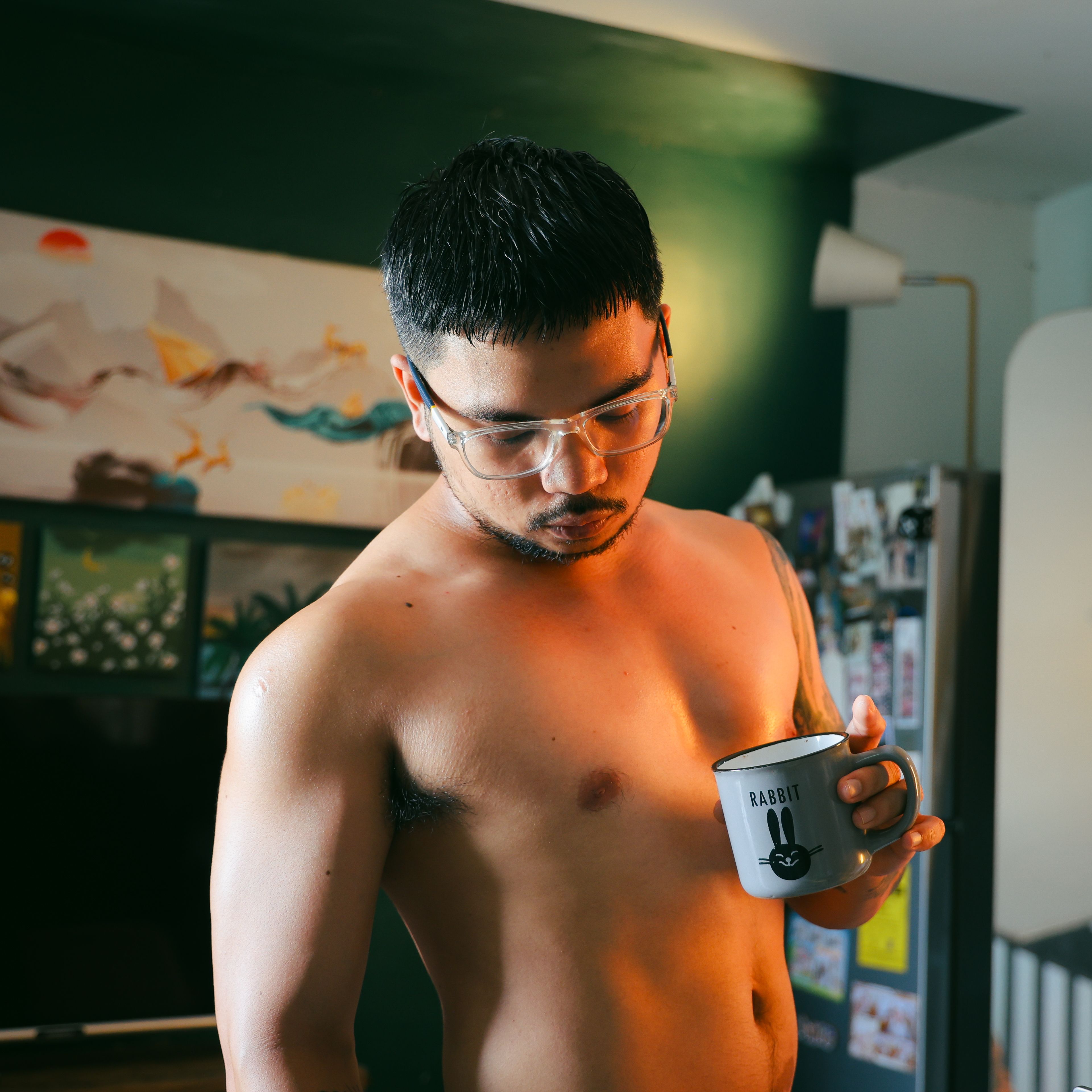 A man with glasses holds a mug, illuminated by warm indoor lighting, displaying a relaxed and thoughtful demeanor.