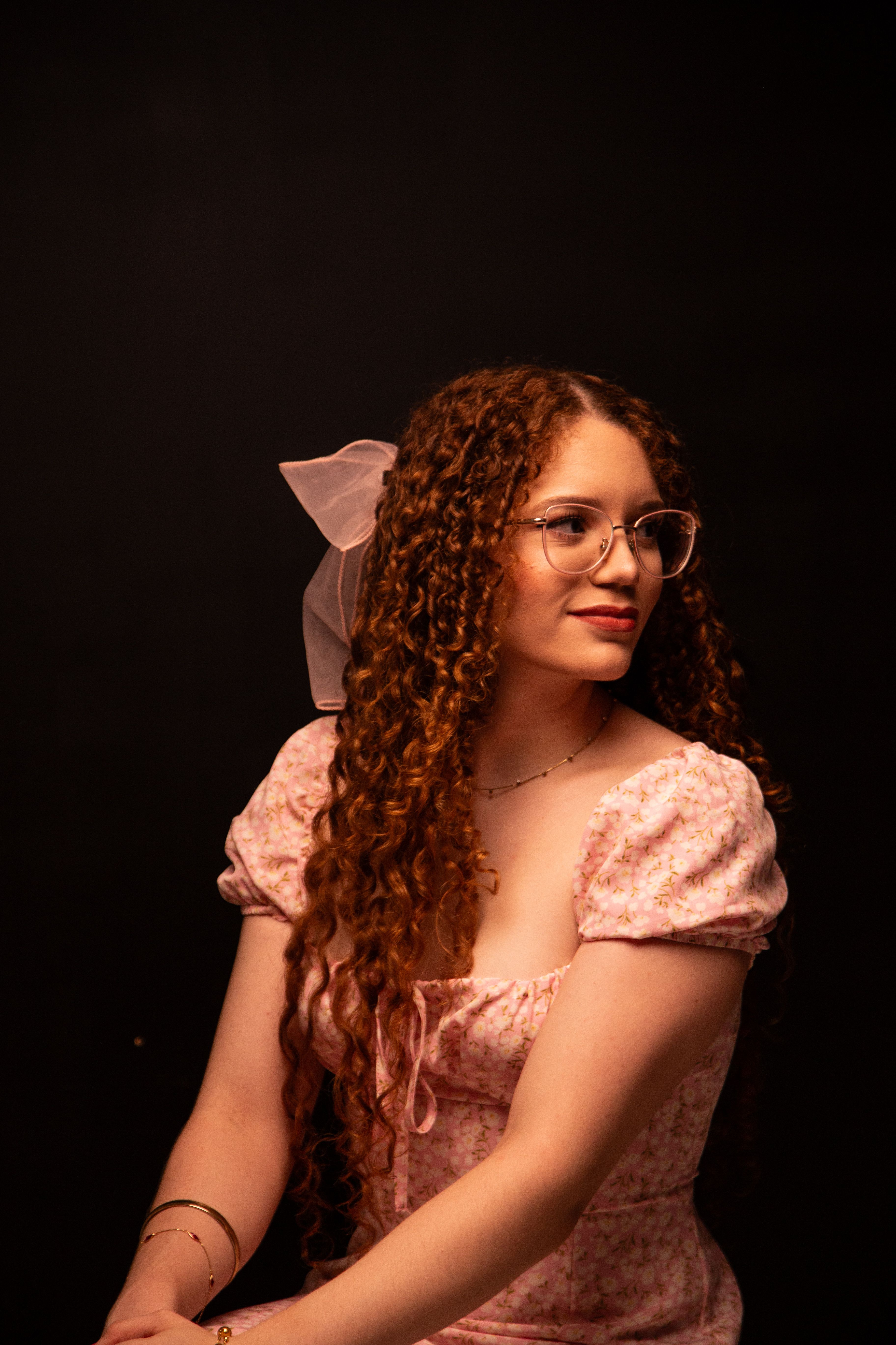 Elegant portrait of a woman with curly hair and glasses, wearing a pink dress against a dark background.