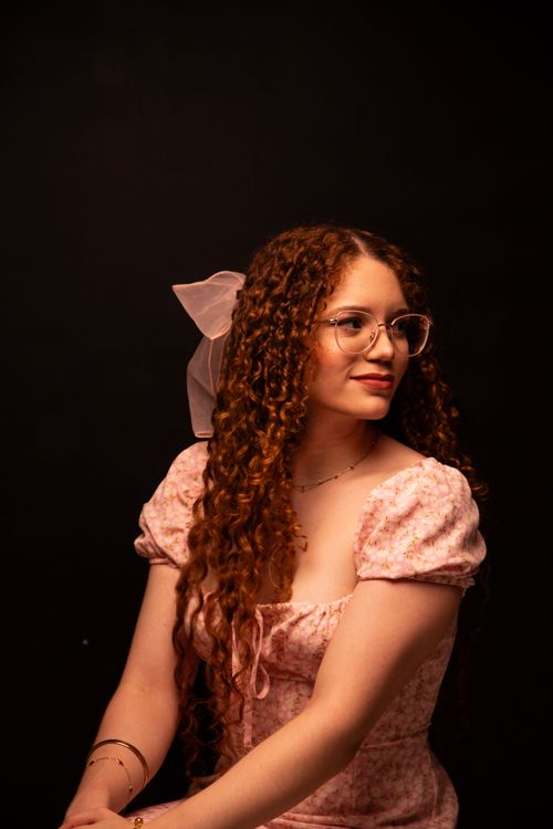 Free Elegant portrait of a woman with curly hair and glasses, wearing a pink dress against a dark background. Stock Photo