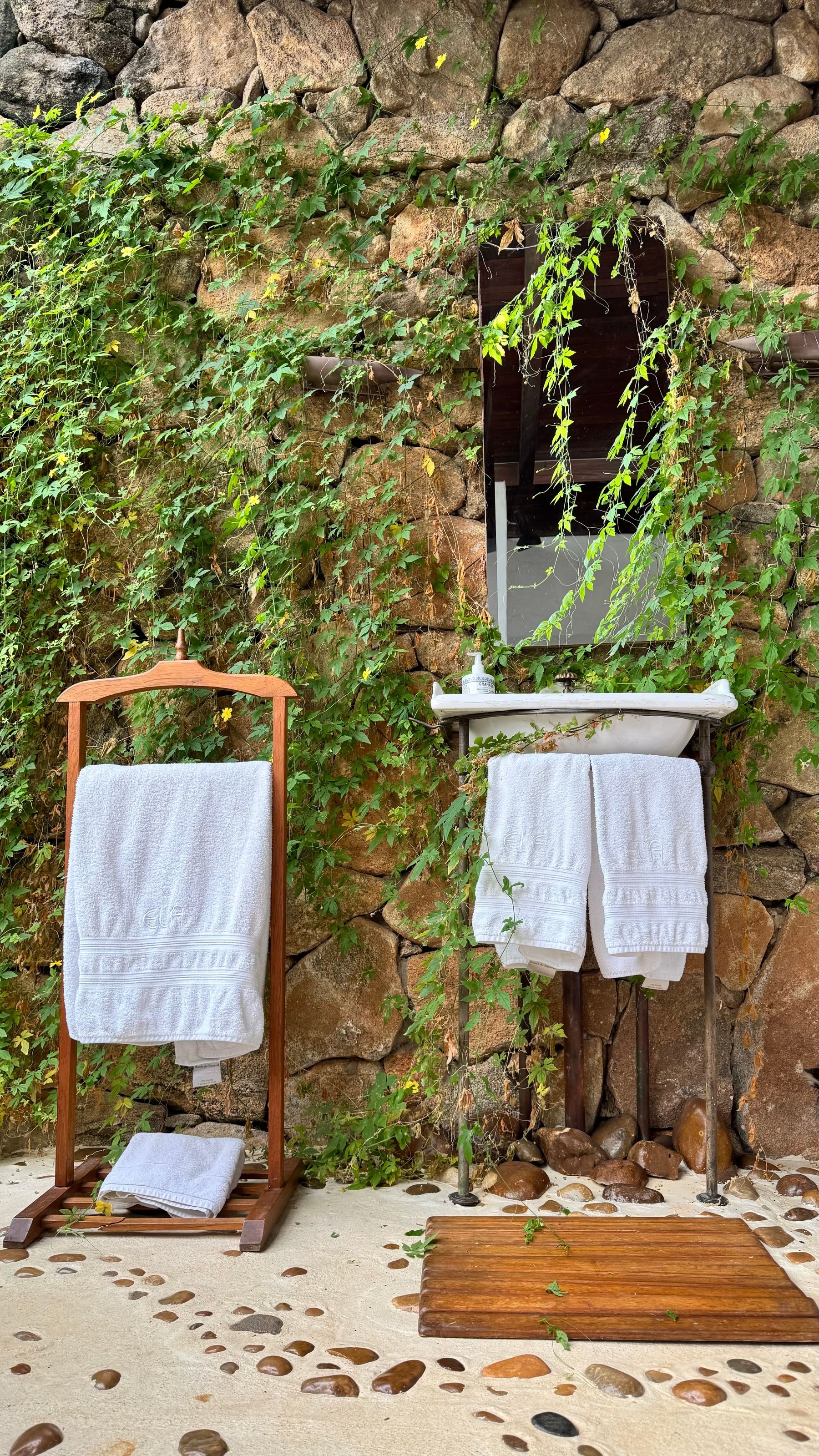 Outdoor bathroom sink and towel stand against a stone wall with vines