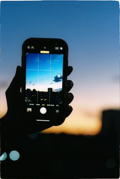 Silhouette of a hand holding a smartphone capturing a city skyline during twilight.