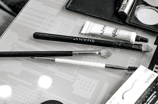 Monochrome image showing makeup brushes and cosmetics on a glass table.