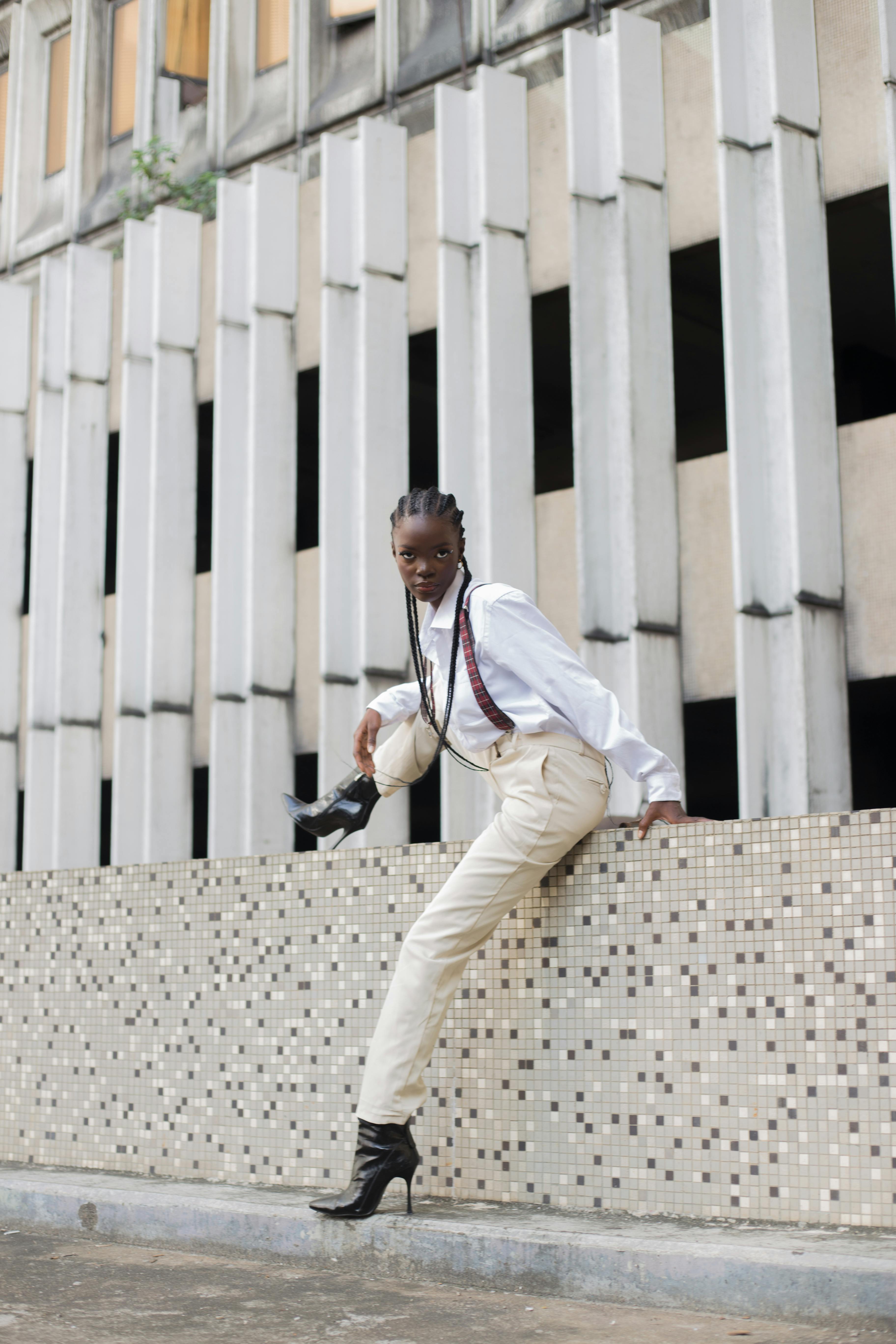 Fashionable woman posing confidently on urban building ledge wearing stylish outfit and boots.