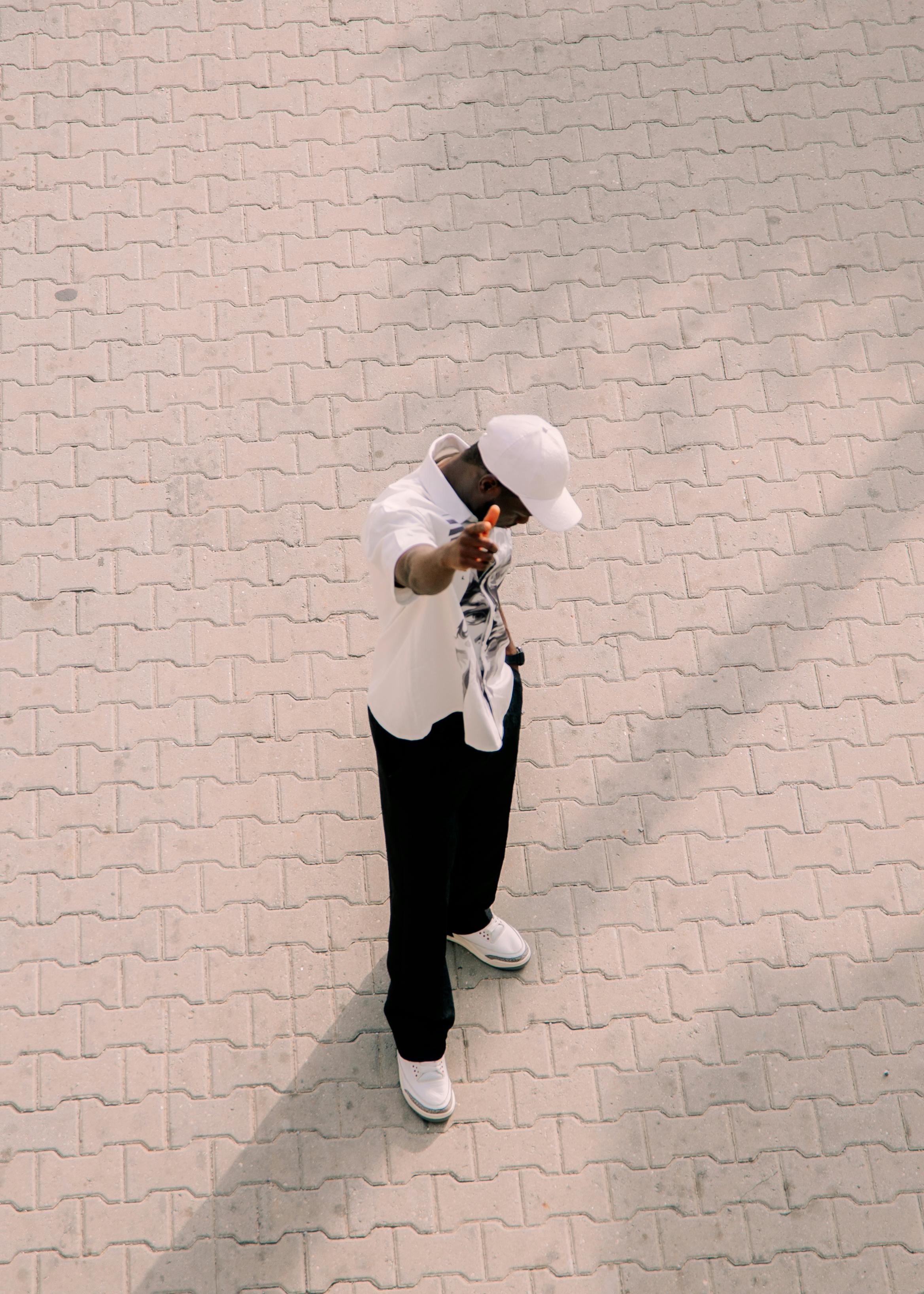 grátis Indivíduo estiloso com roupa branca e boné, visto de cima em uma calçada iluminada pelo sol. Foto profissional