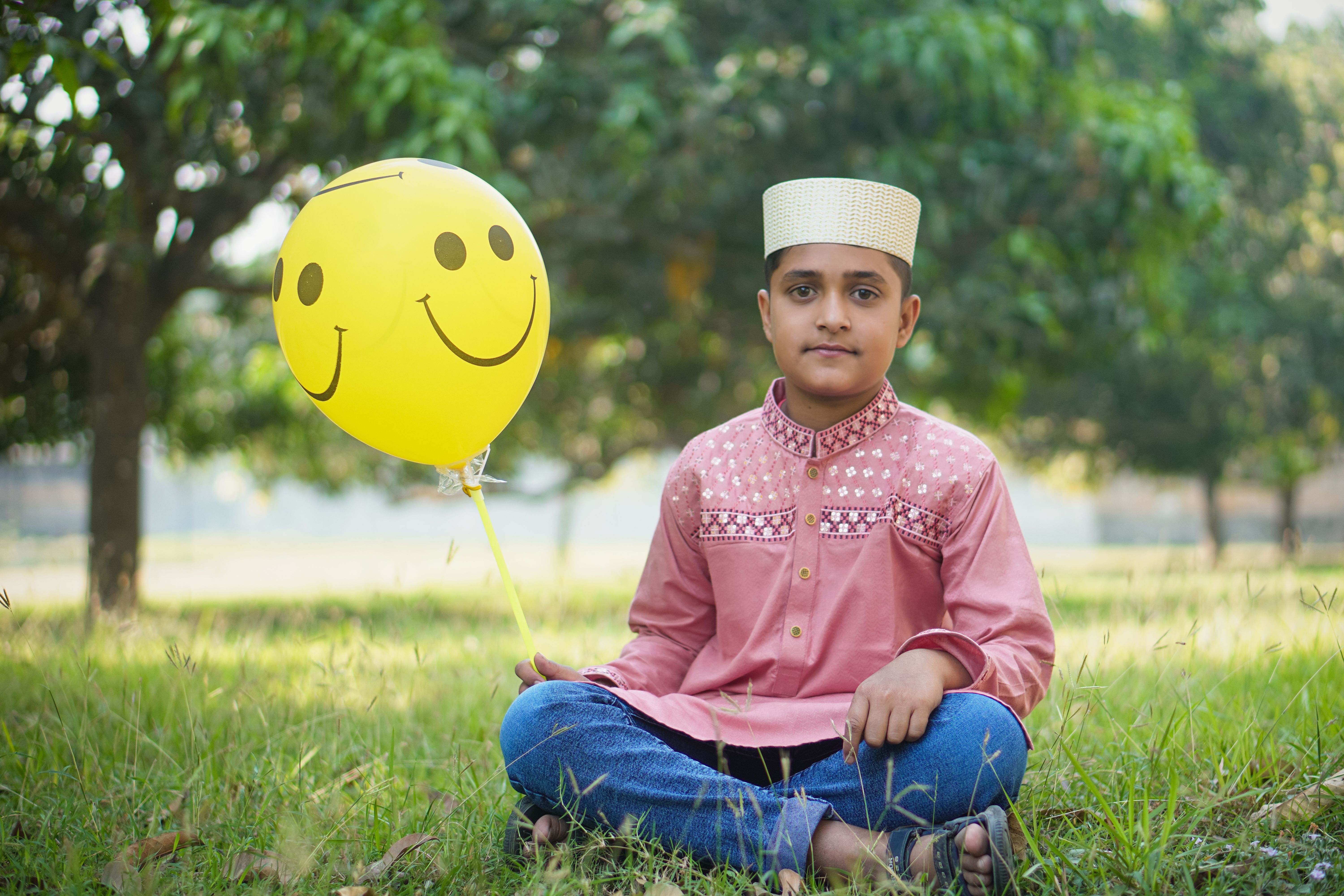 A cheerful child in traditional attire sits outdoors holding a smiley balloon amidst a lush green park.