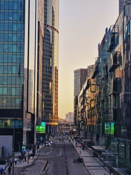 A bustling city street at twilight flanked by towering modern skyscrapers with reflective glass facades.