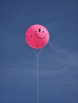 A vibrant pink balloon with a smiley face floating against a clear blue sky.