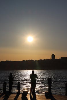 Two silhouetted fishermen casting lines at sunset by a waterfront.