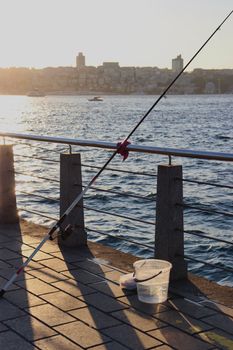 A fishing rod and bucket by the waterfront during sunset with a cityscape in the background.
