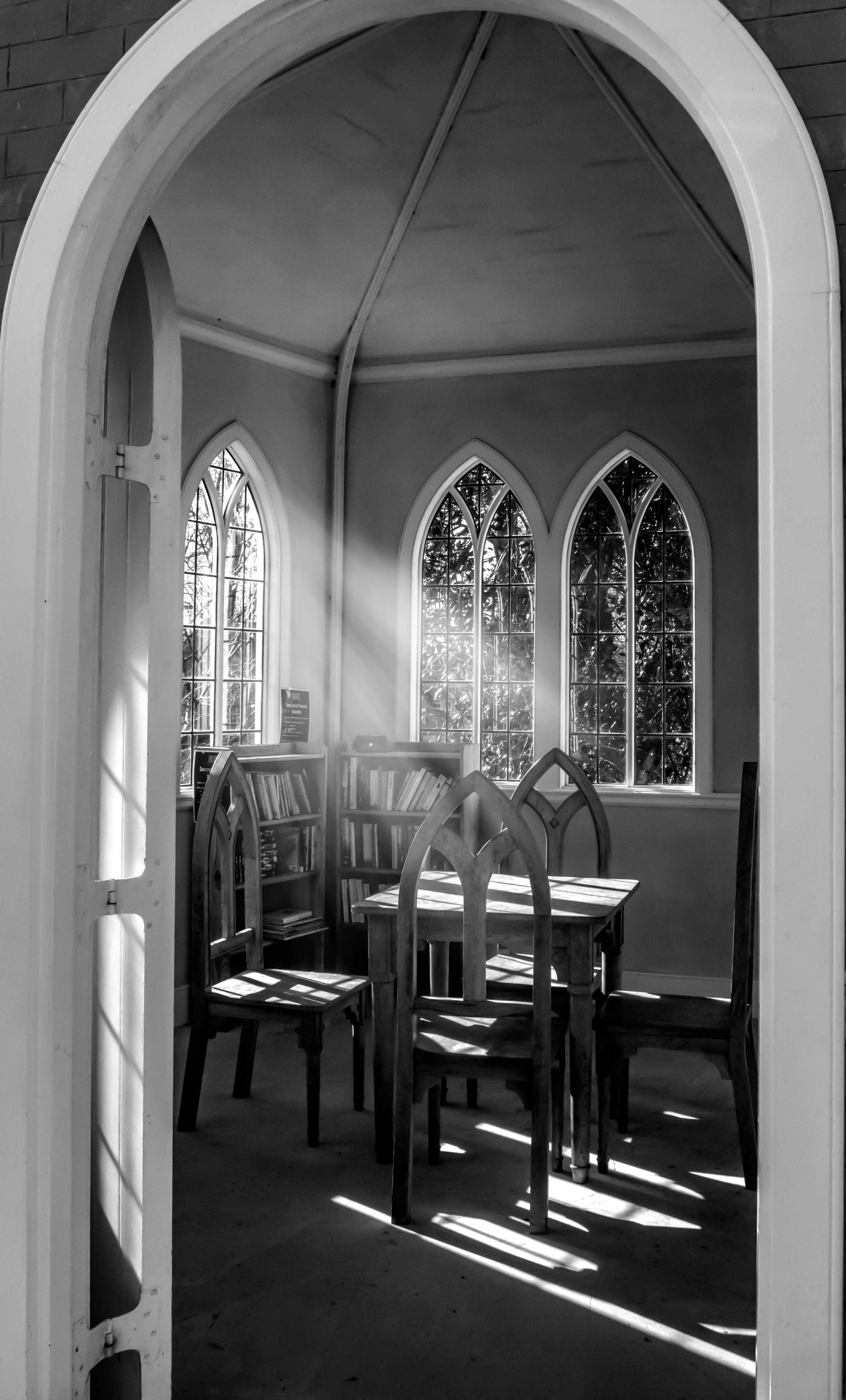 Monochrome view of a peaceful, sunlit room with gothic arched windows and wooden chairs.