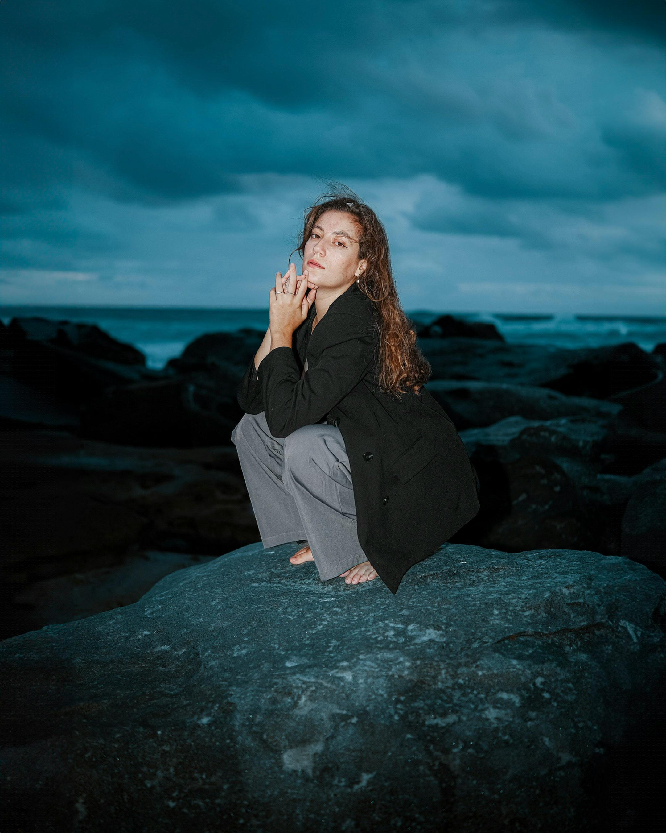 A contemplative woman sits barefoot on rocks by the sea under a dramatic overcast sky.