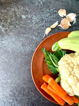 Top view of fresh vegetables including cauliflower, carrots, and garlic on a rustic background.