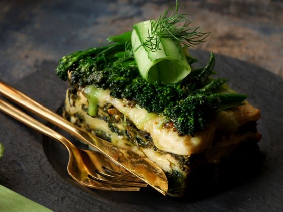 Slice of spinach lasagna topped with broccolini, cucumber ribbon and dill with gold fork