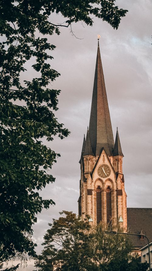 Free A Gothic church spire with a clock and trees at sunset, showcasing architectural beauty. Stock Photo