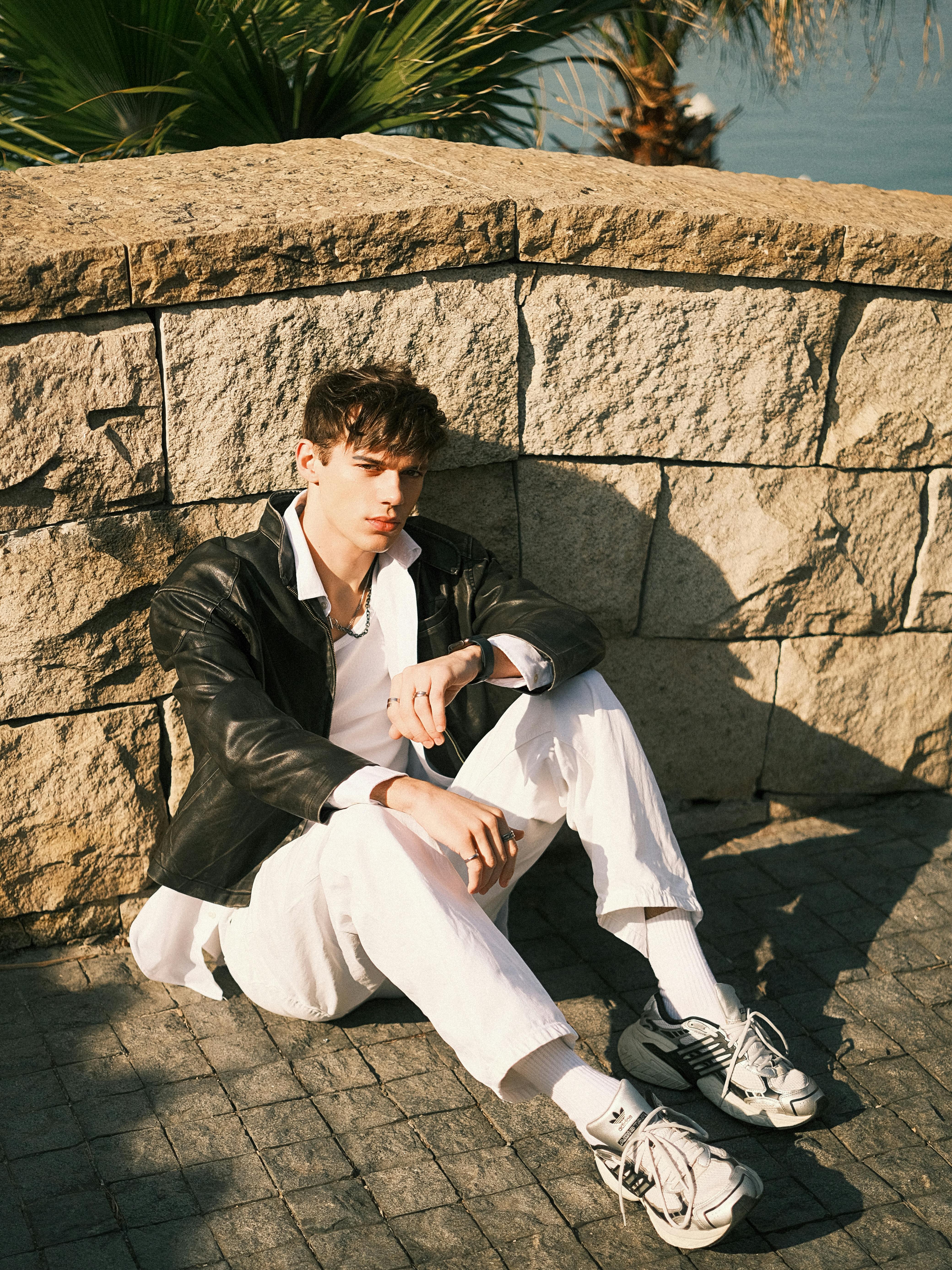 Young man in leather jacket sitting casually against a stone wall, enjoying the sunny day.
