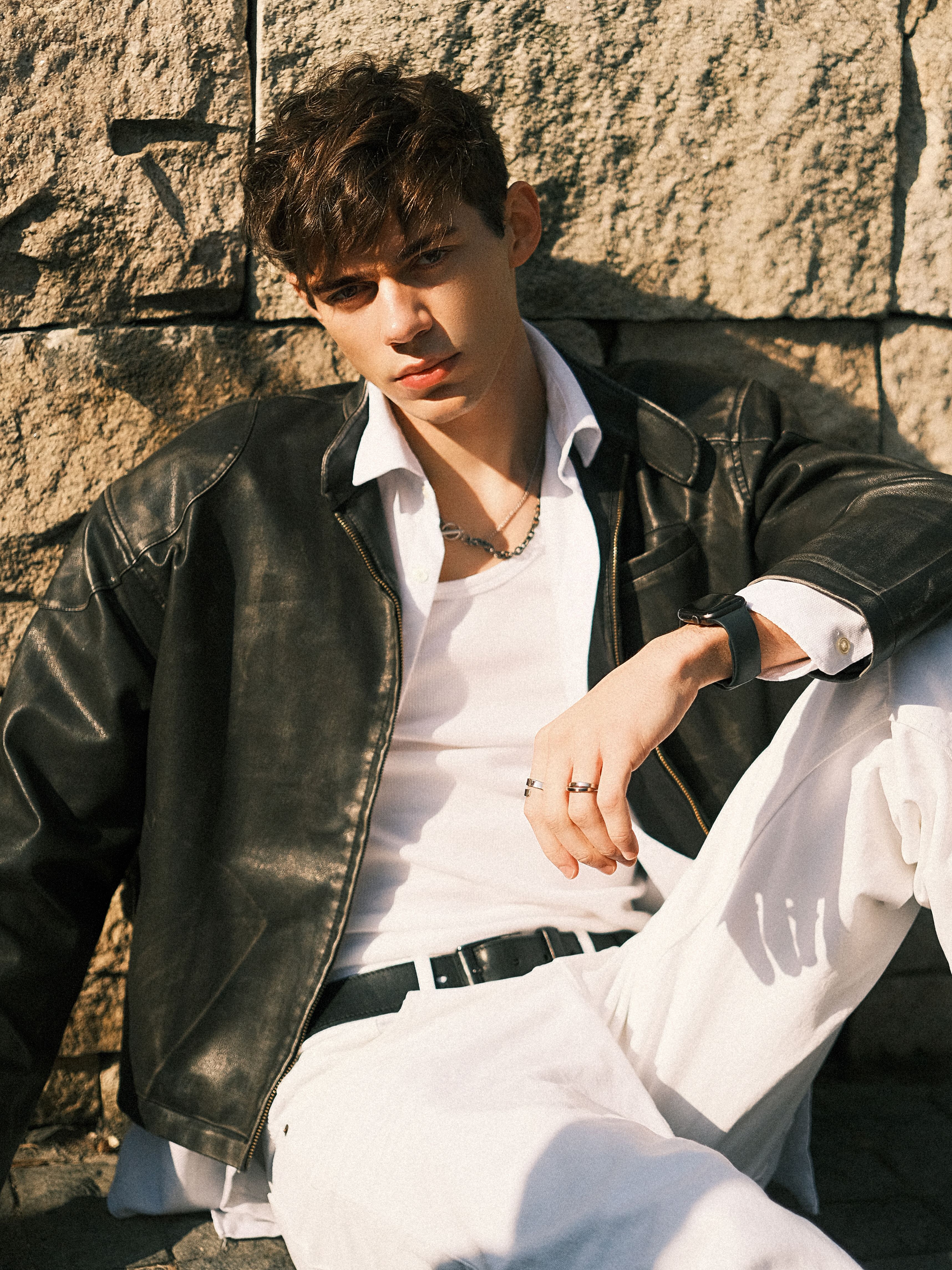 Portrait of a young man in casual attire leaning on a stone wall under the sun.