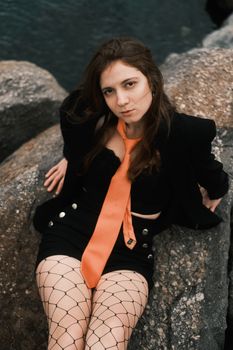 Young woman in fashionable black attire and orange tie, relaxing outdoors on rocky terrain.
