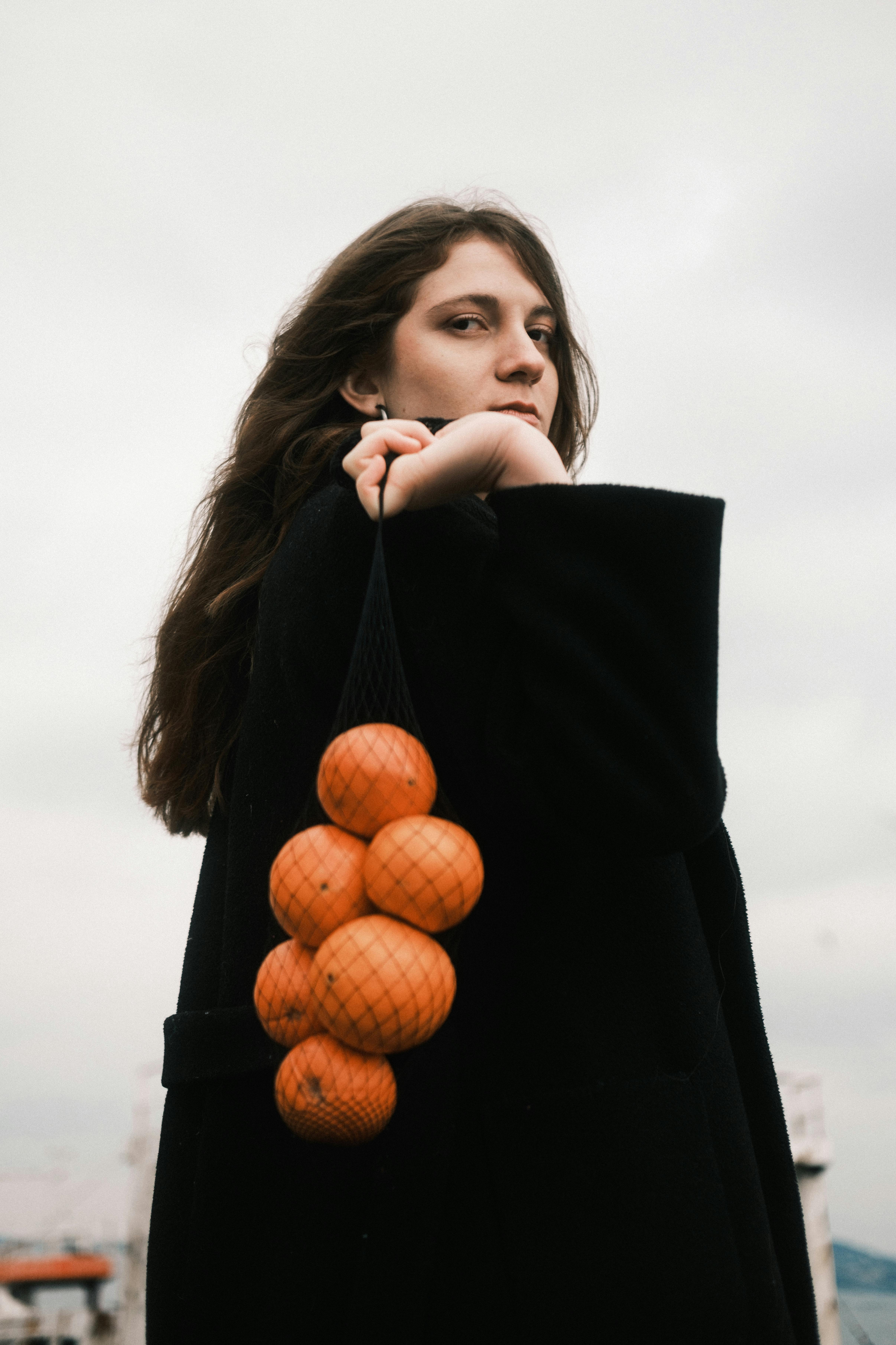 Thoughtful woman in black coat holding oranges in mesh bag against cloudy sky.
