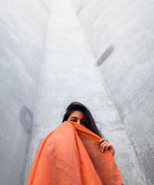 A woman holding an orange scarf in front of a tall urban structure, captured from below.