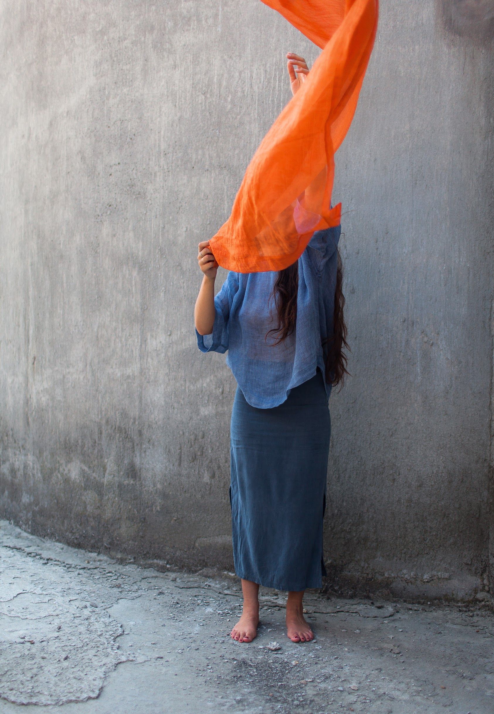 Woman in flowing attire holding orange scarf near a concrete wall.