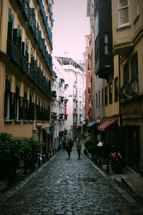 Free Picturesque cobblestone street with historic buildings and pedestrians under a cloudy sky. Stock Photo