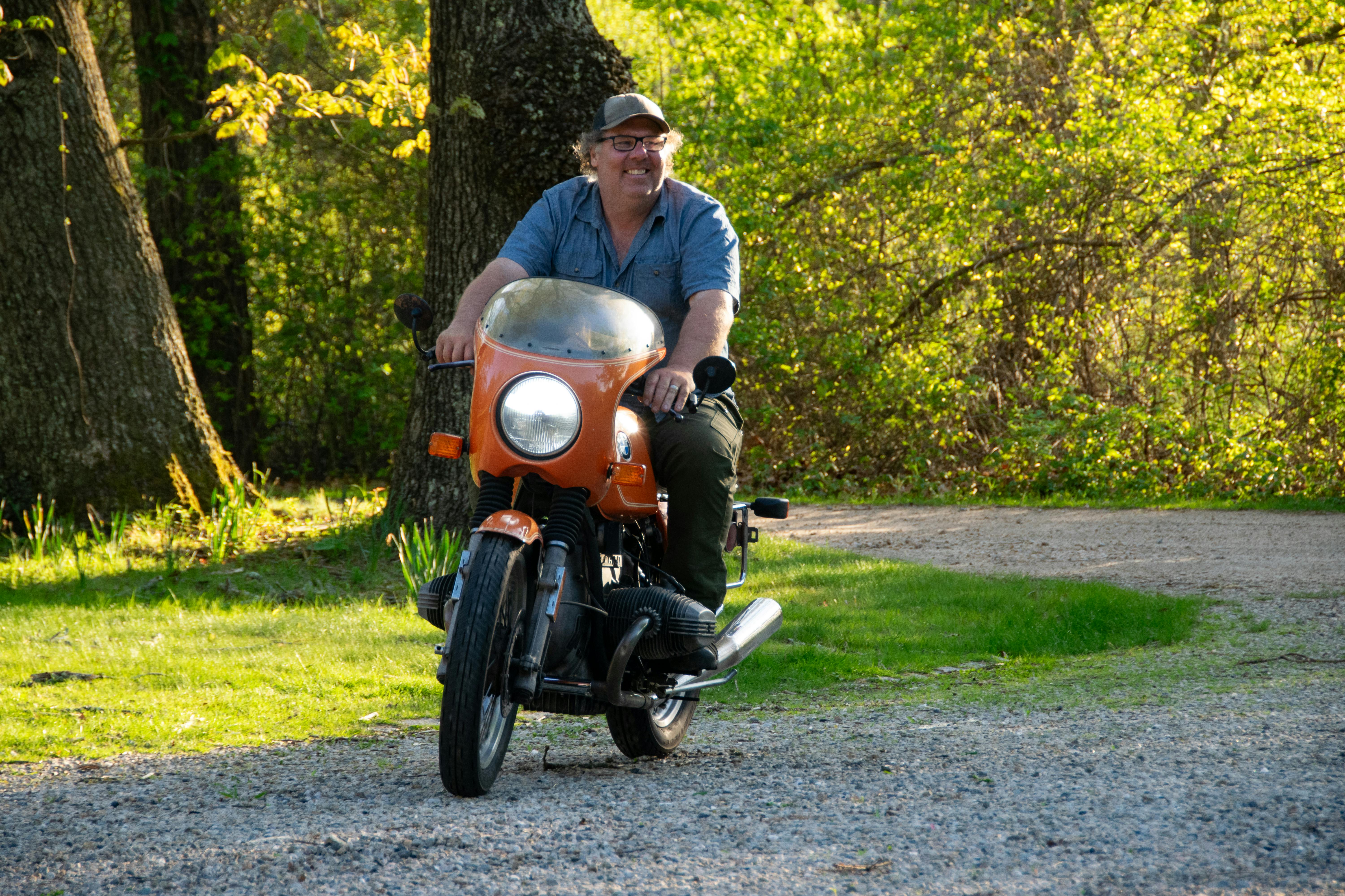 Free Adult male enjoying a ride on a vintage orange motorcycle in a sunlit park setting. Stock Photo