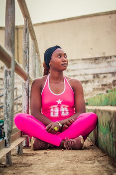 African woman in pink sportswear sitting outdoors on concrete steps, looking thoughtful.