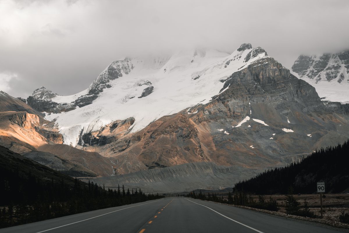 The Icefields Parkway running dead straight toward glaciated peaks, a 90 km/h speed sign on the shoulder, forest on both sides of the road.