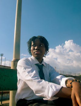 A young adult seated outdoors in formal attire, looking thoughtful against a bright sky.