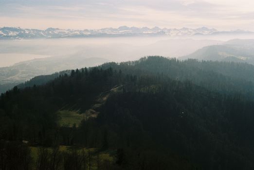 A stunning view of the misty alpine mountains in Switzerland, showcasing layers of forested hills and snow-covered peaks.