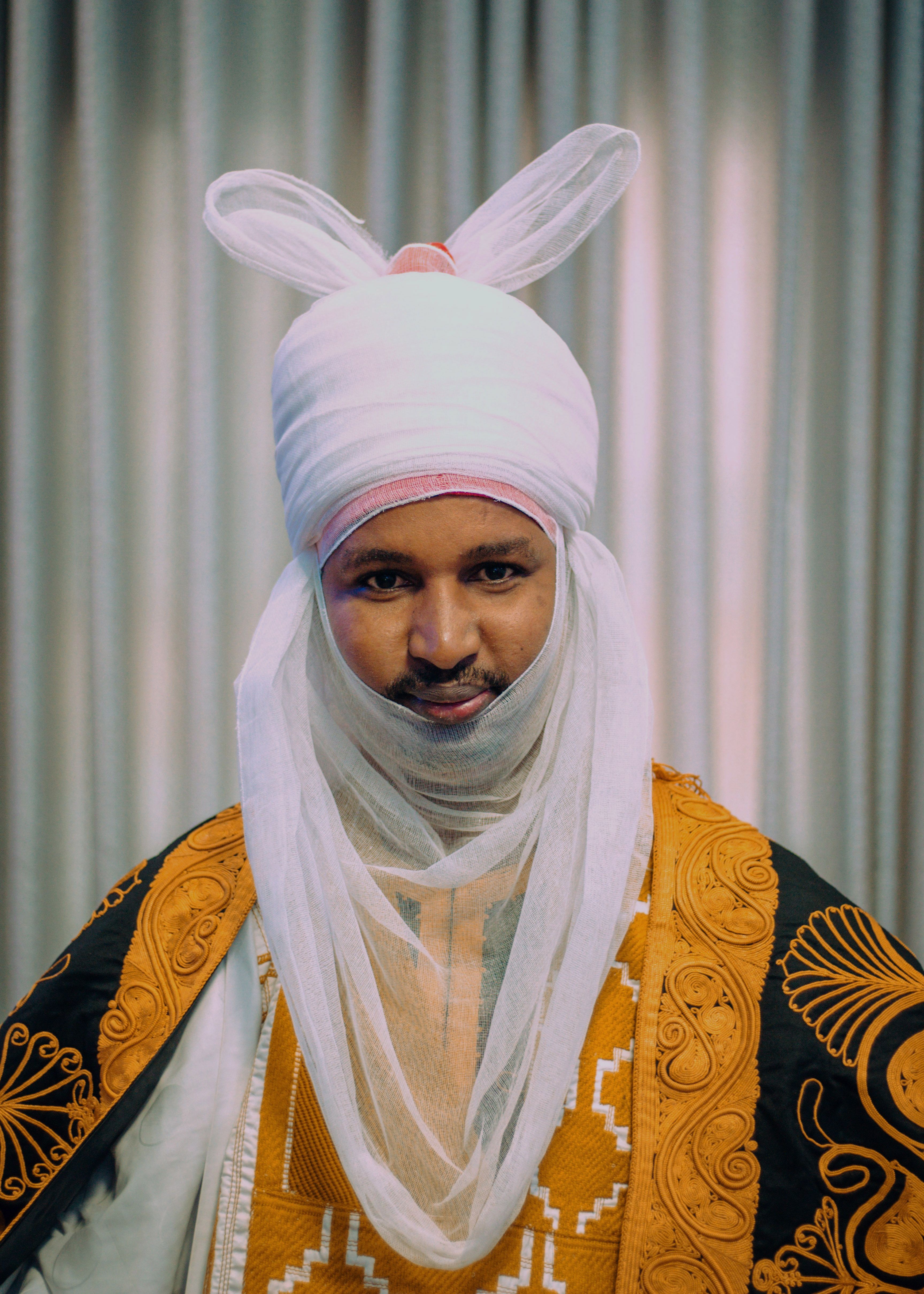 Portrait of a man in traditional African attire with intricate embroidery and a white turban-like headdress.