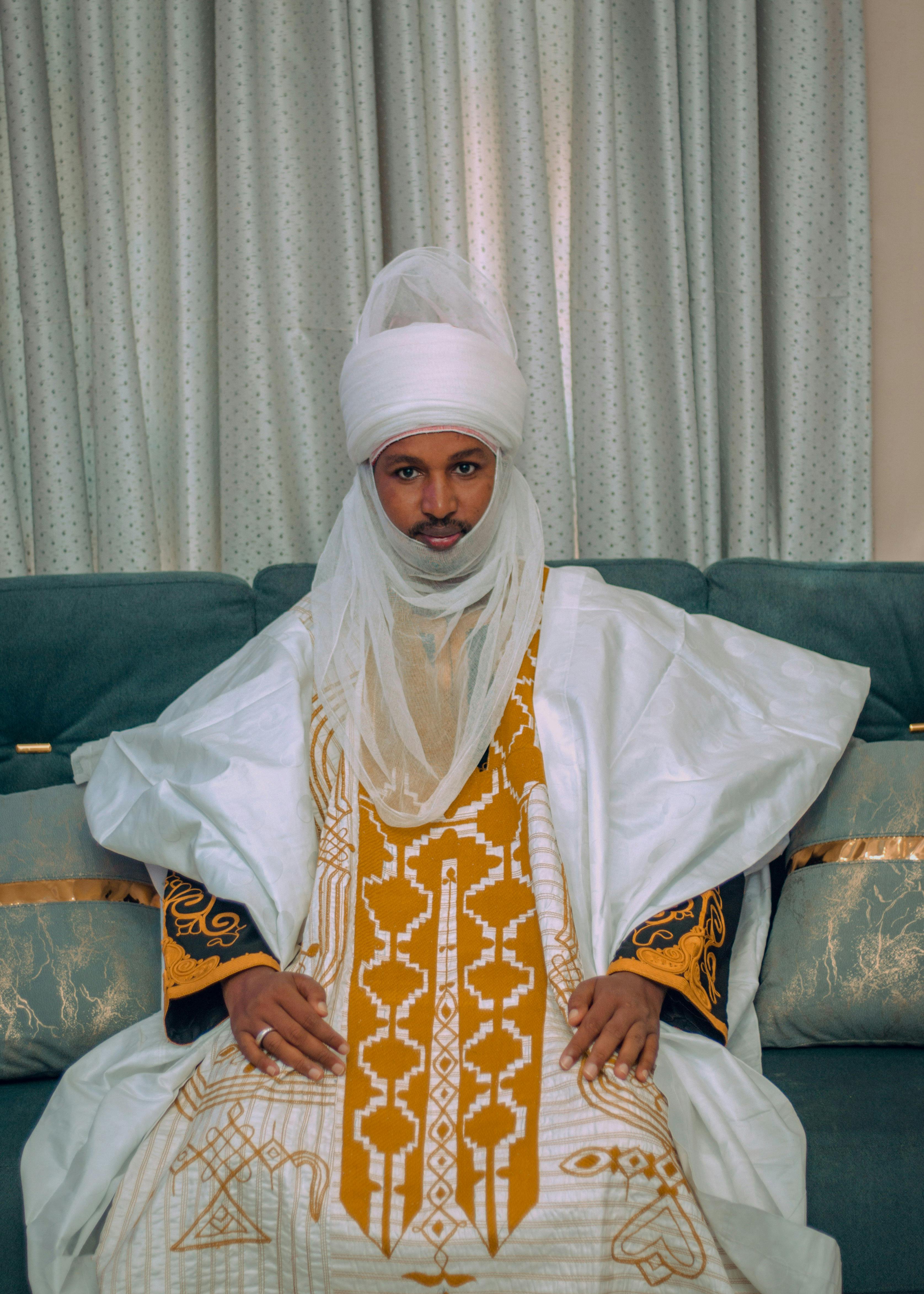 A man in an ornate traditional African robe and turban sits on a couch indoors.