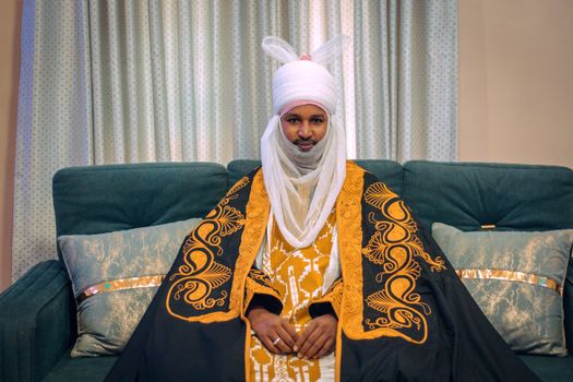 Man seated in traditional African clothing with ornate patterns, indoors.