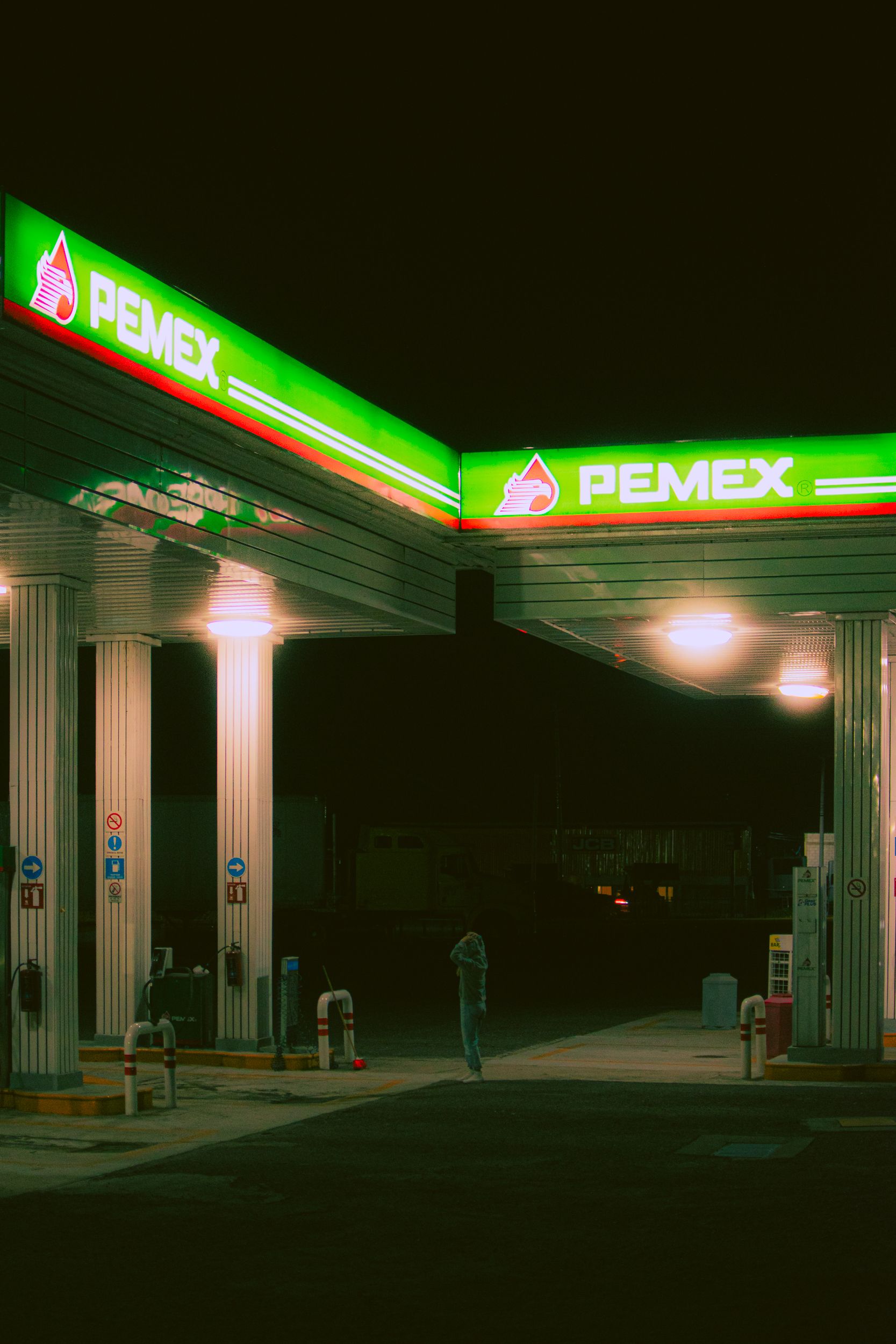 A solitary figure under the illuminated canopy of a Pemex gas station at night, evoking a sense of travel and solitude.