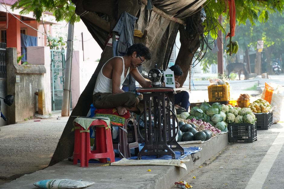 A street tailor repairs clothes beside a vibrant produce market. Urban life intertwined with local commerce.