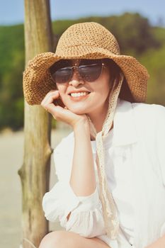 Woman in straw hat and sunglasses sitting outdoors, enjoying a sunny day with a big smile.
