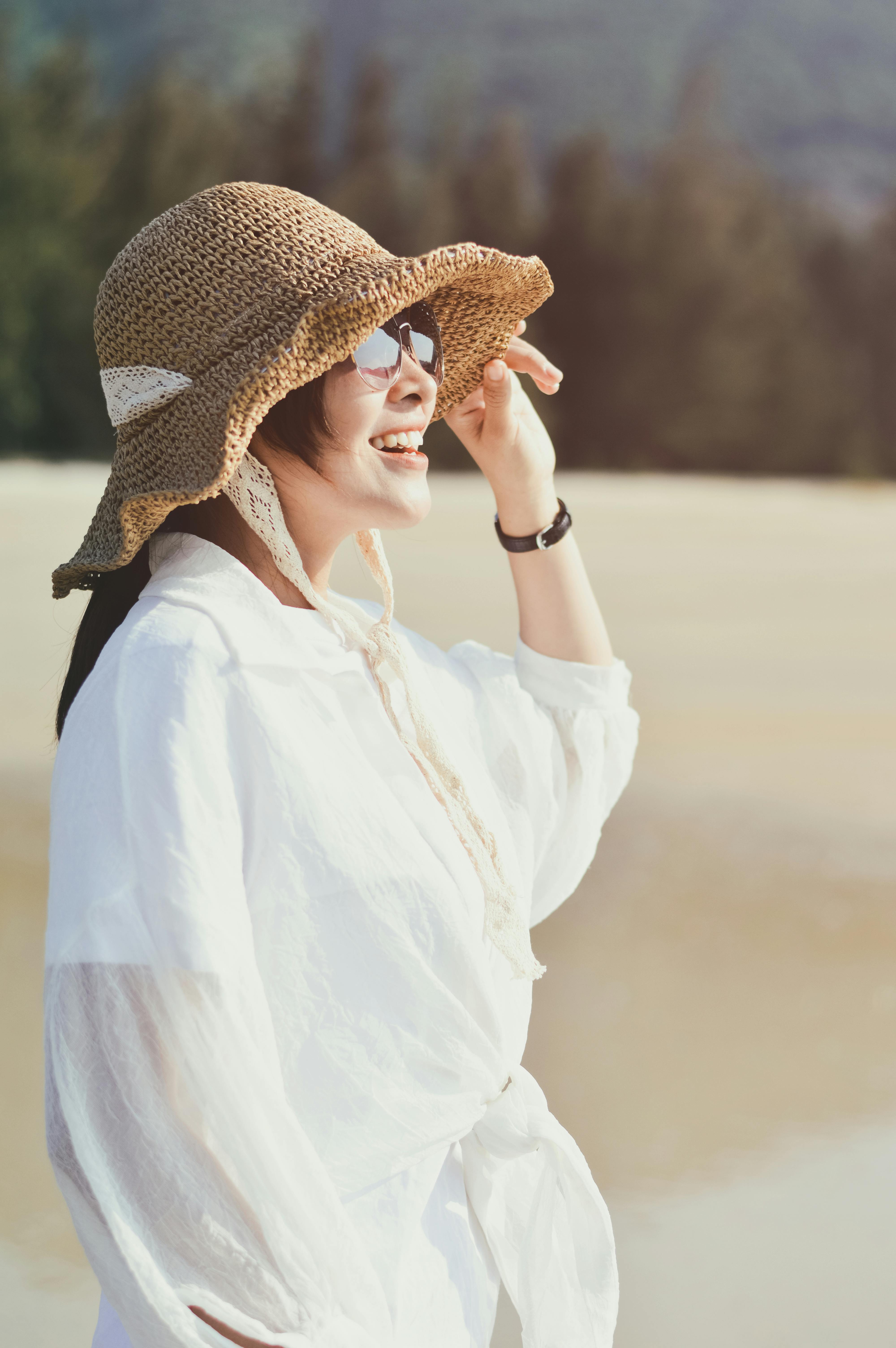 Woman in a sun hat and sunglasses smiling brightly at a sunny beach.