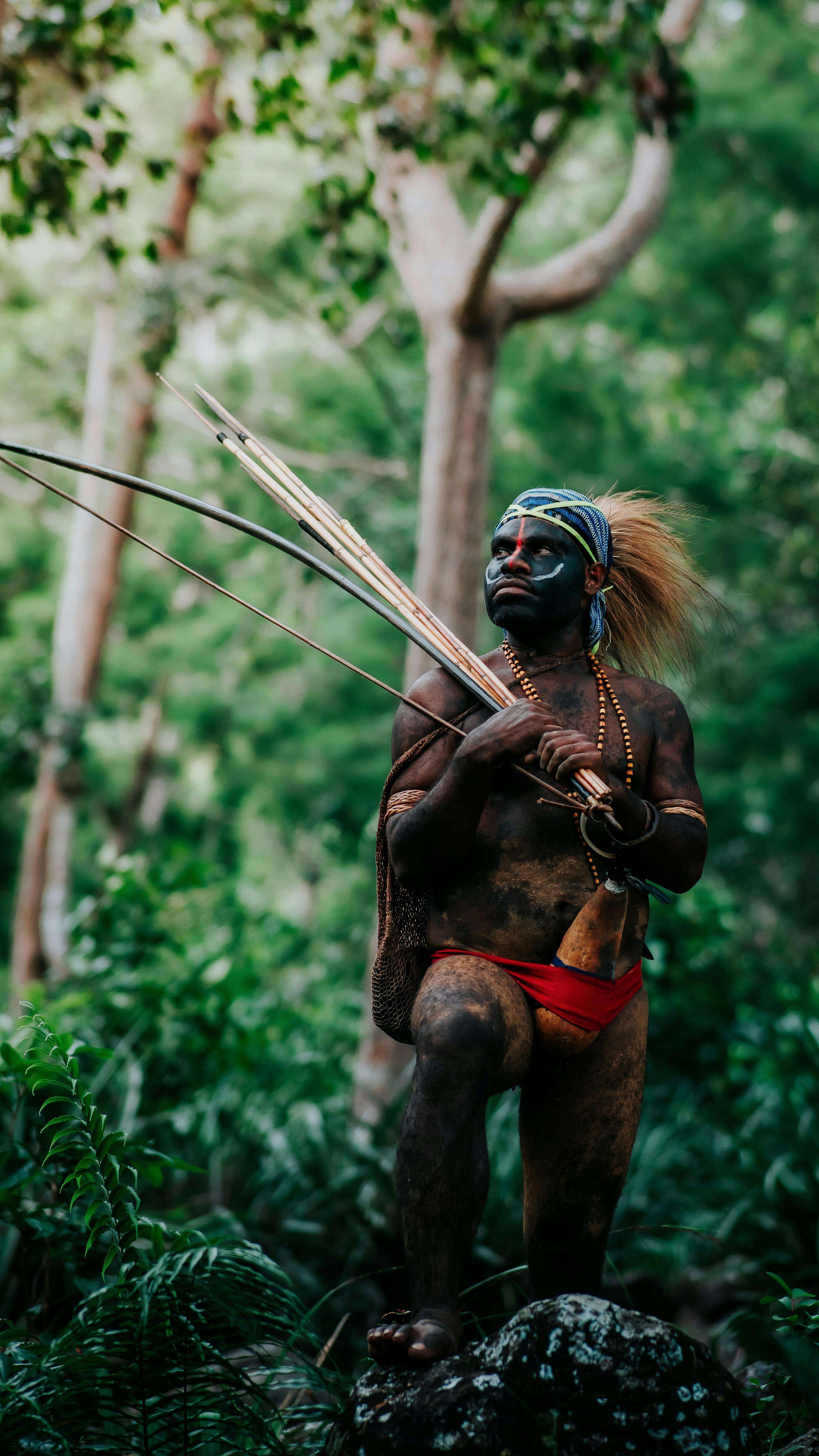 Indigenous person in traditional attire with bow and arrows in a verdant forest.