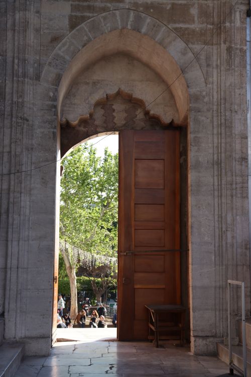 Free Large wooden door at stone archway opens to a lush courtyard with people and trees. Stock Photo