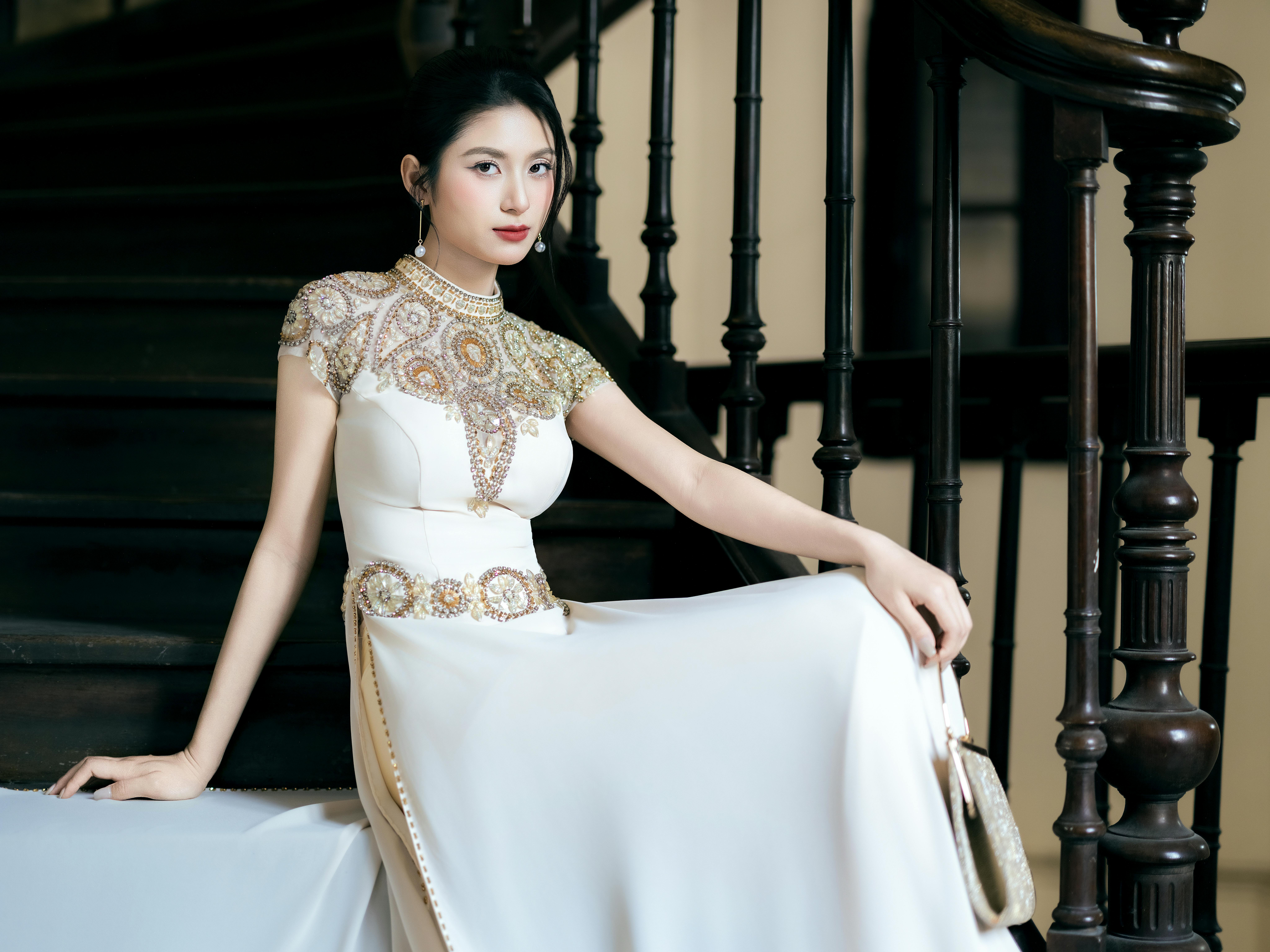 Stylish woman in a white evening gown posing elegantly on a staircase indoors.