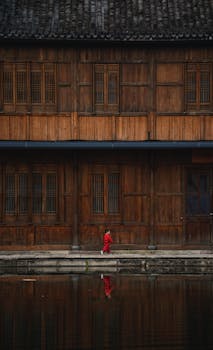 A child in red walks by a traditional wooden building reflected in water, creating a serene, cultural scene.