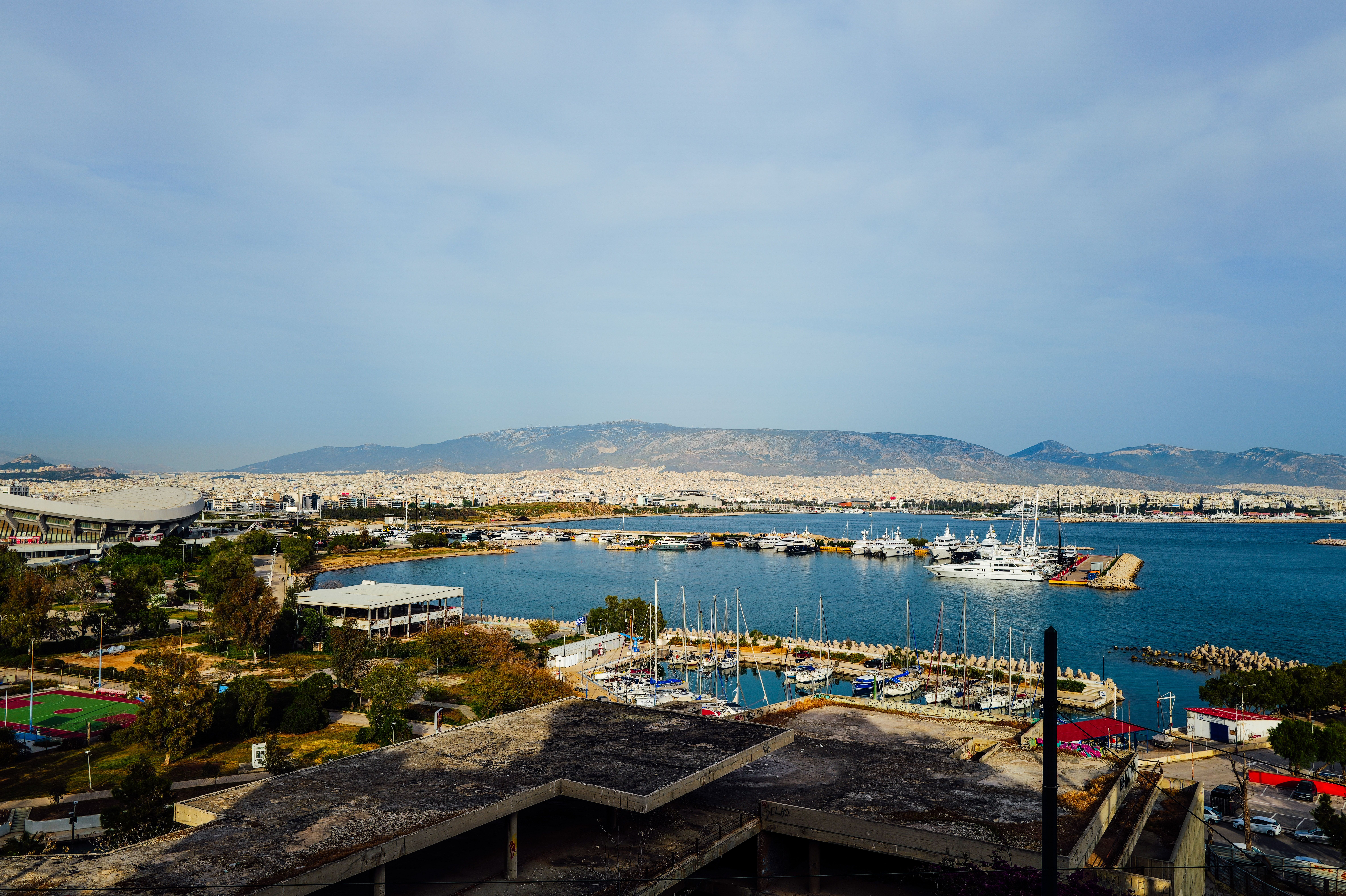 A picturesque marina view with yachts, cityscape, and distant mountains under a clear sky.