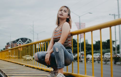 Free A young woman squatting outdoors by a yellow railing in a city setting, expressing casual fashion and confidence. Stock Photo