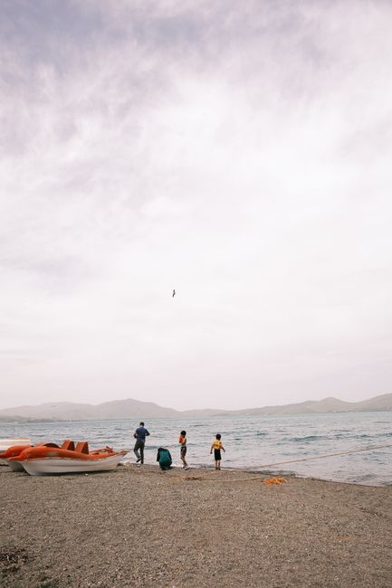 Family on a pebbled beach with boats and sea in the background under a cloudy sky.