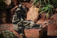 A detailed bronze statue depicting two children seated on a bench in a garden setting, surrounded by rocks and plants.